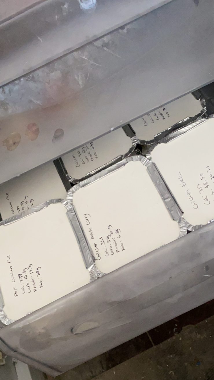 Photo of several small silver foil trays with white lids in a clear freezer drawer. The white lids have various meals and nutrition information written on them.