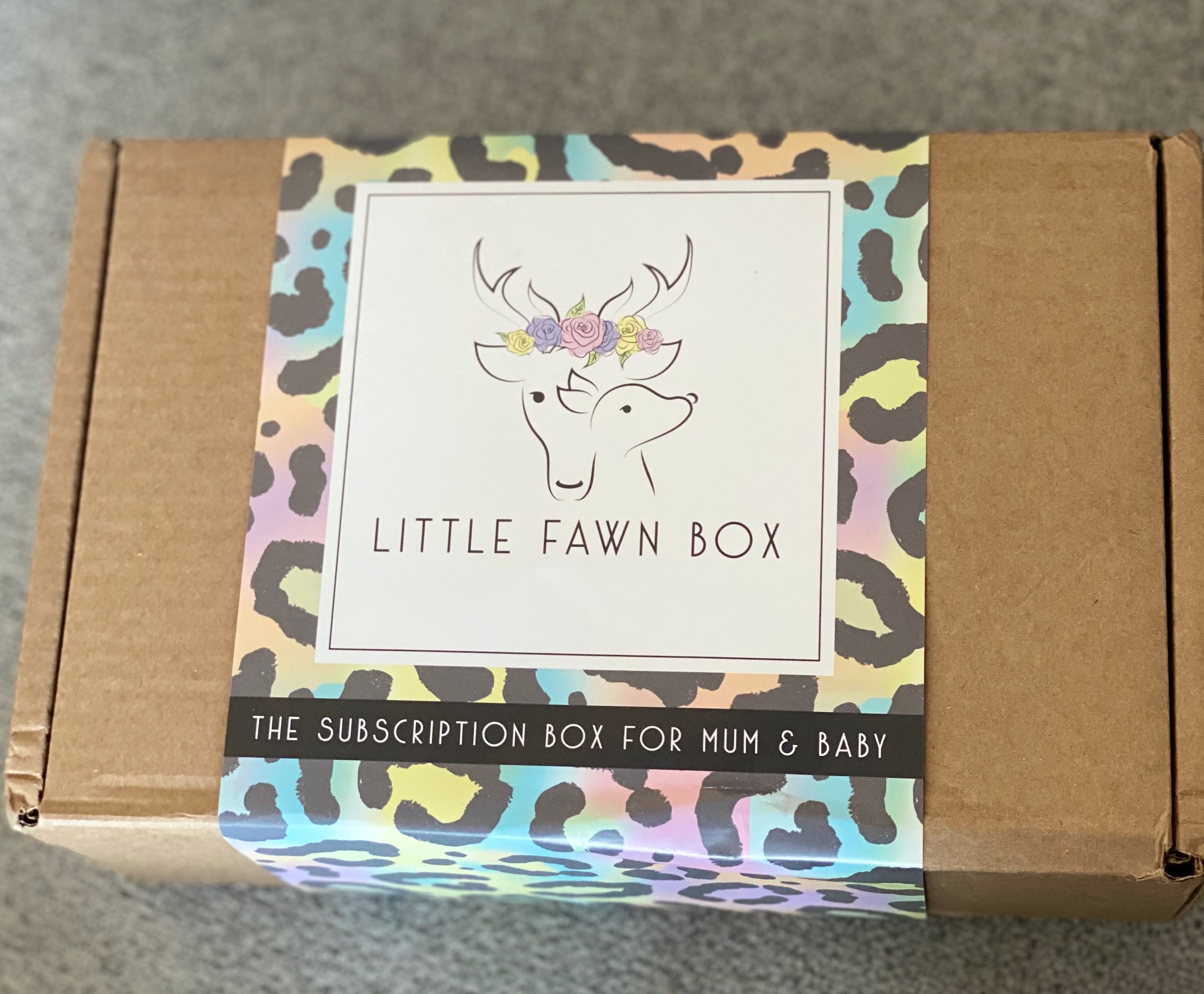 Photo of a brown box on a grey background with a multicoloured animal print sleeve wrapped around it. The sleeve also has a cream coloured square box with a drawing of a dears head with flowers one, and a fawn just in front of it. The words 'little fawn box' are in black underneath. There's also the words 'the subscription box for mum & baby' in cream on a black background underneath.  
