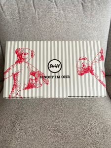 Photo of a grey and white striped box with drawings of teddy bears in red on. In the middle of the box is a cream circle with black outline which has the word Steiff in block writing in. The words knopf 