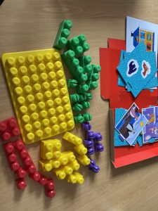 Bright yellow, green, red and purple building blocks are laid out on the floor. To the right is an open box containing several pieces of the playmat in with various colours. 