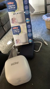 Photo of the Nuby dummy Steriliser and box in the background. There is also a black battery charger and white USB cable in the photo. 