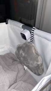 Photo of a white Blink Mini Camera with black face mounted on a white and grey Snuzpod bedside crib. There is also a grey Snuz cloud attached to the Snuzpod in the photo. 