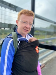 Alex, a white man with red hair and wearing glasses is looking towards the camera smiling. Alex is wearing a blue and pink jacket, and a black baby wrap which has Alice, a small white baby with brown hair inside. 