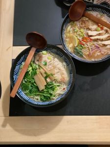 2 blue and white bowls with brown wooden ladles on the side of them are sitting on a light brown wooden table with black quartz marble rectangles on the table top. The bowls are filled with two different types of ramen. 