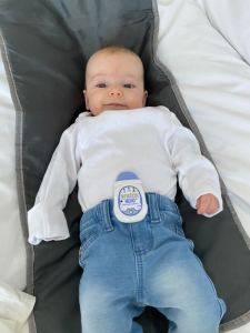 Alice, a small white baby with brown hair is looking at the camera with eyes open wearing a white long sleeved vest and light blue jeans. On Alice's jeans is a white and purple Snuza Hero MD breathing monitor.