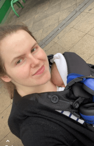 a selfie Steph, a white woman with brown and blonde hair wearing a black baby carrier with Alice, a small baby with brown hair and wearing a white jacket inside. The baby carrier has blue and blue ear defenders and a blue Chelsea bib attached to it. 