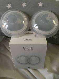 A photo of two clear plastic Elvie Catch collection cups with silicone backing sat on top of a white box which they come in. The box has a photo of the collection cups on it along with the words Elvie Catch in grey on top of the picture. 