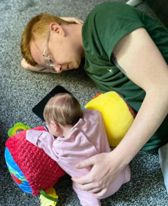 Alice a small white baby with brown her is wearing a pink sleepsuit whilst leaning on a red and yellow tummy time pillow. She is being supported on it by Alex, a white man with red hair and glasses, wearing a dark green t-shirt. 