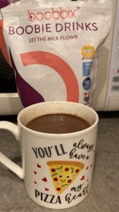 A photo of a white mug with a slice of pizza on and the words ,you'll always have a pizza my heart, in black writing on the front containing hot chocolate is on a grey kitchen worktop. There is a white, pink and purple bag behind the mug with the words 'boobbix boobie drinks let the milk flow' on. 