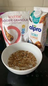 Image shows a white bowl with granola and a milk alternative in on a black worktop. Behind the bowl is a white, pink and purple bag with an image of granola on and the words 'boobbix boobie granola let the milk flow' on. Next to the bag is a white Alpro coconut and almond milk container with images of almonds and  coconut on. 