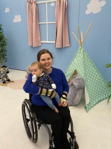 Steph, a white woman with her brown and blonde hair in a ponytail is sat in her wheelchair wearing a blue jumper, black trousers and hand splints whilst holding Alice, a white baby with brown hair who is wearing a dark blue floral top with light blue jeans. Behind them is a green tent with a large grey cuddly toy by it. Behind on the wall is a false window with pink curtains. 
