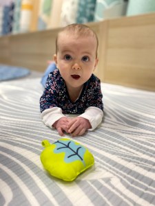Alice, a white baby with brown hair wearing a dark blue long sleeved top with floral patterning on is laying on her tummy whilst holing her head up on a grey and cream striped play mat. In front of Alice is a bright green toy with a blue tree on. 