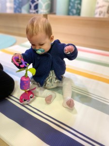 Alice, a white baby with brown hair is sat playing on a multicoloured striped patterned play mat with a pink, purple and green rattle which has a character on at the bottom. Alice is wearing a dark blue jacket, white tights with pink detailing on the heel and toes and a multicoloured dress. Alice also has a blue coloured soother in her mouth.