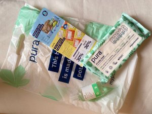 Photo of the contents of the Pura goodie bag. In the photo is a white plastic bag with light green leaf pattern on, with Pura written in dark blue. There is also some additional text in white on a dark blue background. On top of the bag is a bottle of hand sanitiser, a leaflet with discounts on and a pack of Pura baby wipes in light green and cream packaging with darker green leaves on. 