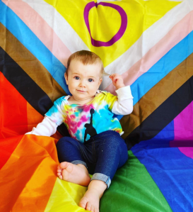 Alice, a white baby with brown hair is wearing a rainbow coloured vest with white long sleeved vest underneath and dark blue jeans. She is sat on an Intersex-Inclusive Pride flag and holding her hair in one of her hands.