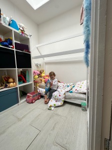 Alice, a small white toddler with dark blonde hair is wearing a white long sleeved top and blue jeggings. Alice is sat on a toddler house bed reading a book. 