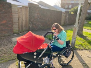 Steph, a white woman with brown hair is outside in the sunshine wearing a green floral wrap top and blue jeans. Steph is sat in her grey and black wheelchair whilst holding onto the handlebar of a red, grey and black pushchair which is in carrycot mode. 