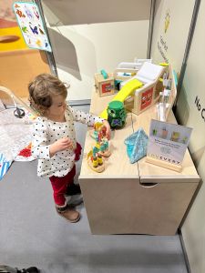 Alice, a small white toddler with dark blonde hair wearing a white and black polkadot top and red trousers stands at a wooden table with several colourful wooden toys on. She is playing with the wooden boats. The photo is taken from above. 

