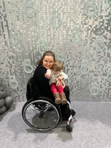 Steph, a white intersex woman with brown hair is wearing a black dress and black cardigan. She is sat in her silver and black wheelchair with Alice, a small white toddler with dark blonde hair wearing a white and black polkadot top and red trousers sat on her lap.