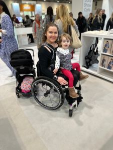 Steph, a white intersex woman with brown hair is wearing a black dress and black cardigan. She is sat in her silver and black wheelchair with Alice, a small white toddler with dark blonde hair wearing a white and black polkadot top and red trousers sat on her lap. Around Alice and Steph is an animal print themed carrier on with all the buckles at the front. 
